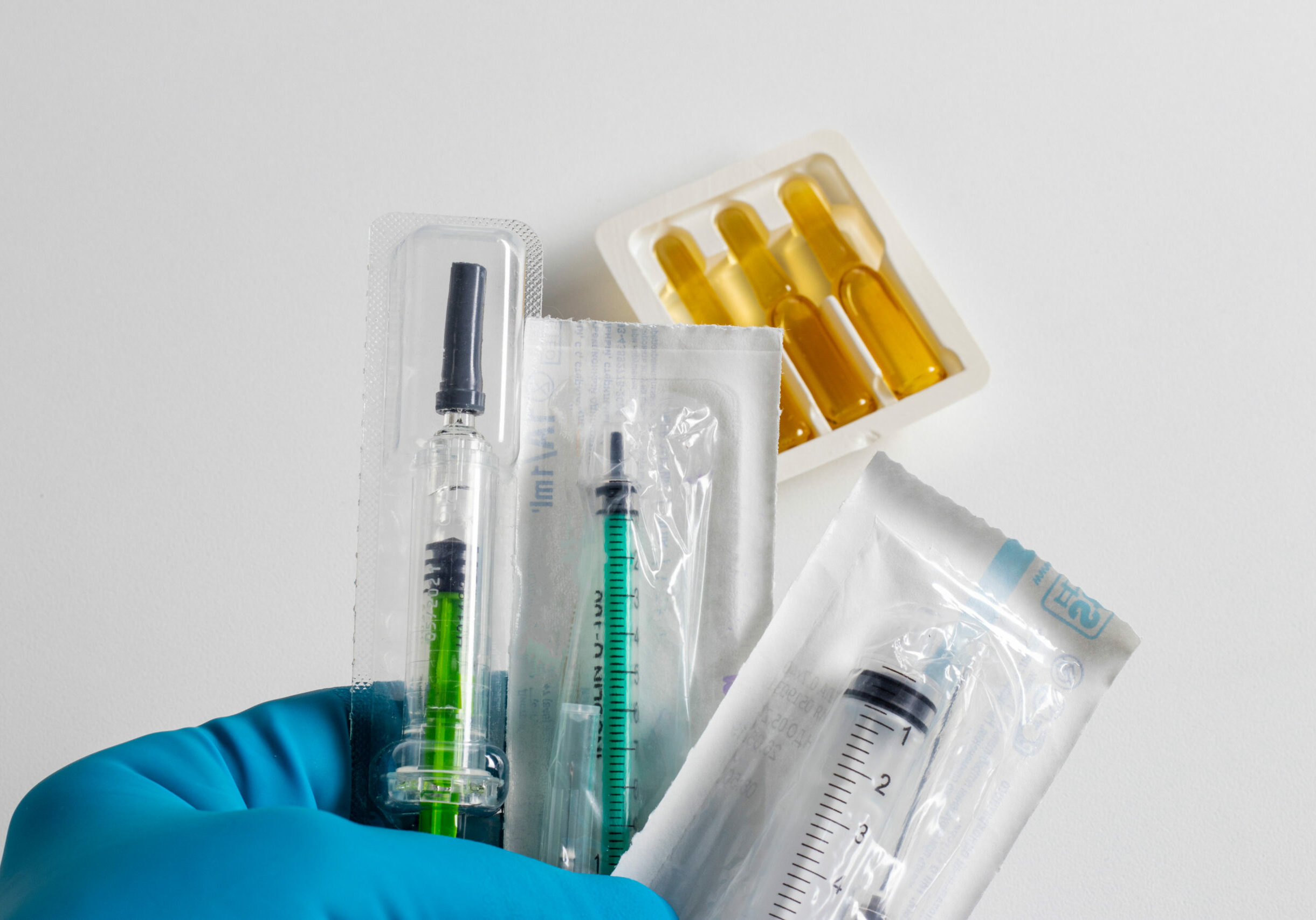 A hand in a medical glove holders syringes, ampoules in the background.